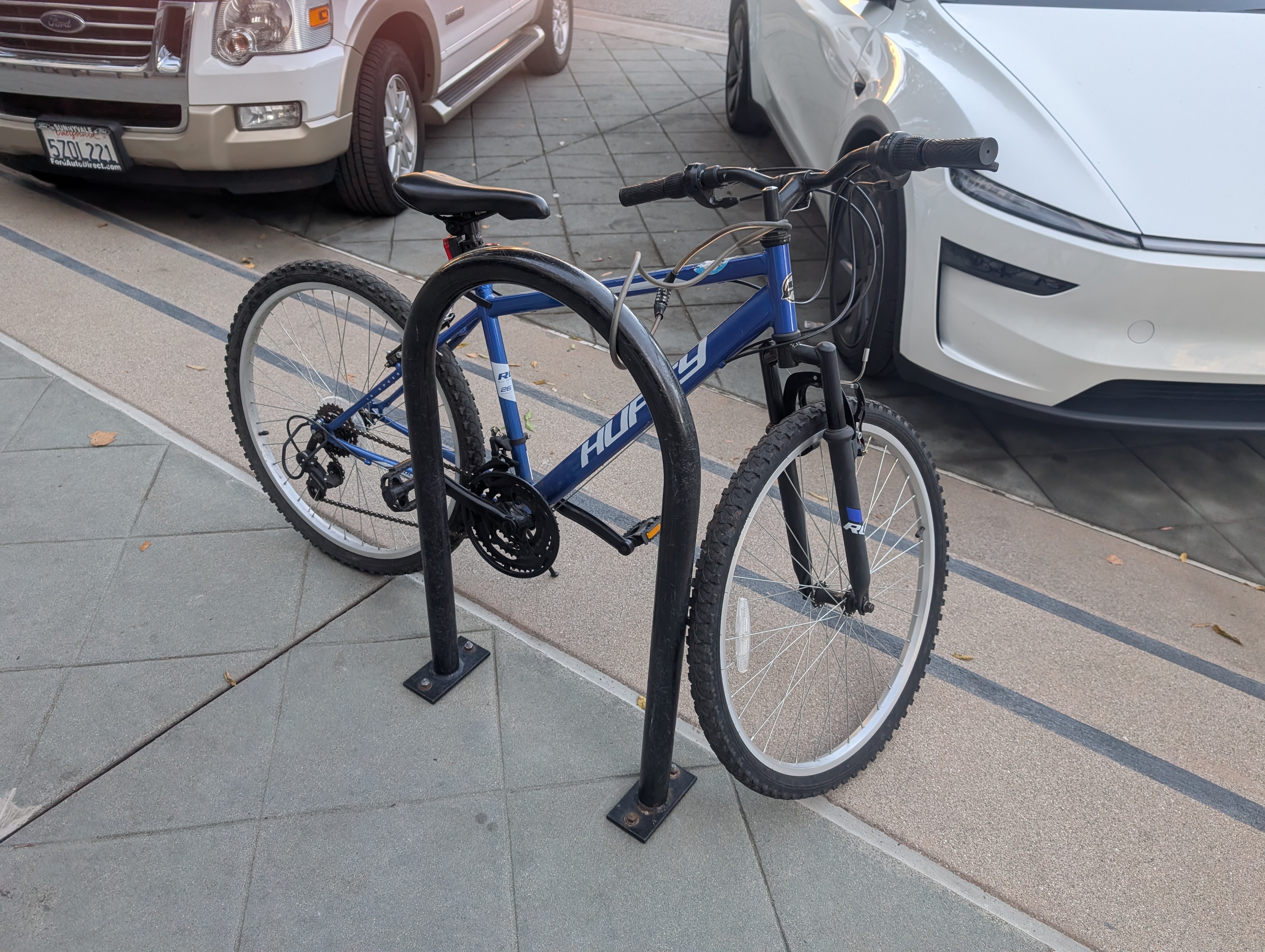 A blue Huffy bike next to a U-shaped bike rack. There is a thin cable combination lock wrapped around the handlebar stem and the bike rack.
